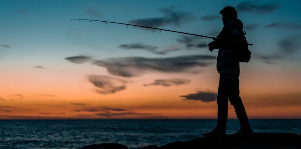 man fishing on shore with sunset in back