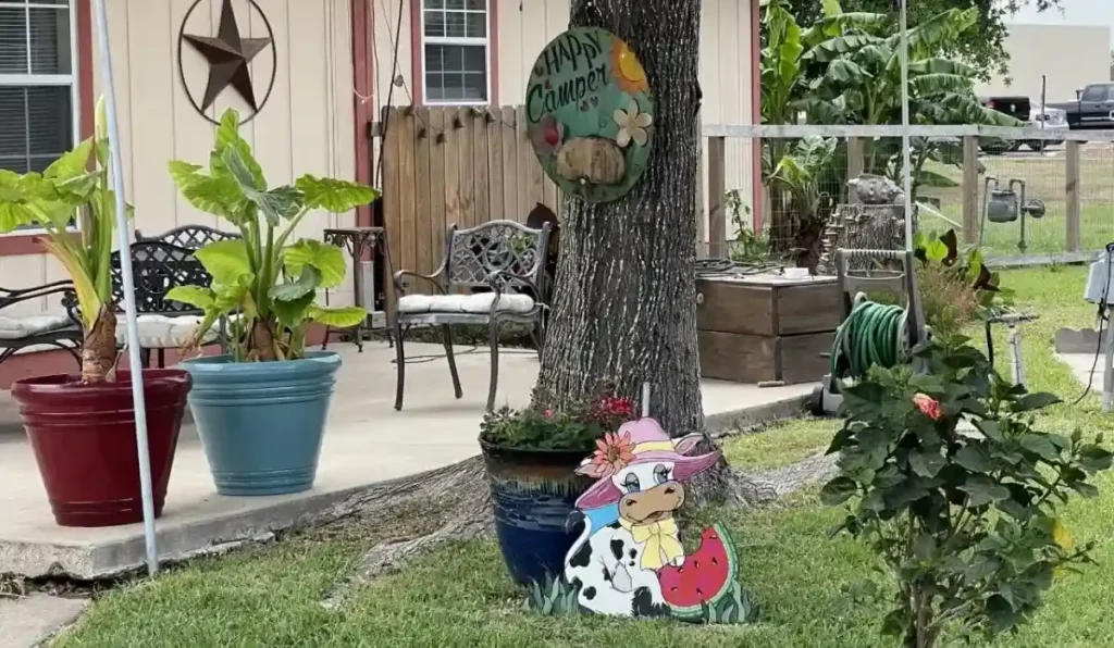 A colorful outdoor scene with a 'Happy Camper' sign, a tree, potted plants, and a cartoon cow cutout.